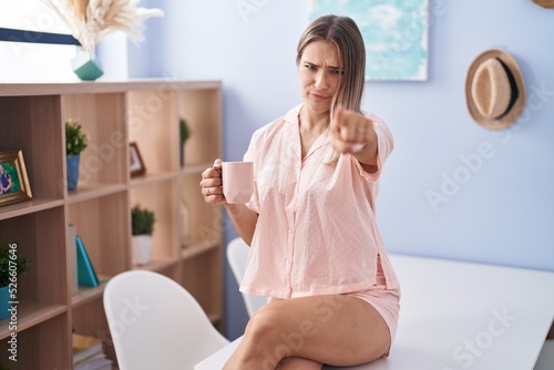 Blonde caucasian woman drinking a cup coffee wearing pajama pointing with finger to the camera and to you, confident gesture looking serious
