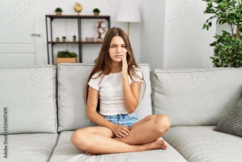 Young brunette teenager sitting on the sofa at home touching mouth with hand with painful expression because of toothache or dental illness on teeth. dentist