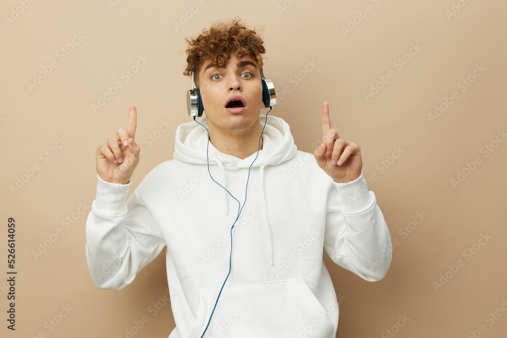 a cute, attractive man stands on a beige background in a white hoodie on his head, listening to music with headphones and pointing his index finger up, mouth wide open a cute, attractive man stands on a beige background in a white hoodie on his head, listening to music with headphones and pointing his index finger up, mouth wide open