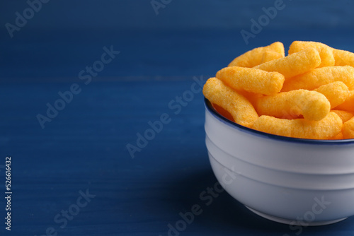 Bowl of tasty cheesy corn puffs on blue wooden table, space for text
