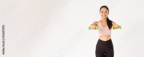 Sport, wellbeing and active lifestyle concept. Cheerful smiling asian fitness girl, sportswoman lifting dummbells, workout on muscles, gaining biceps with home exercises, white background photo