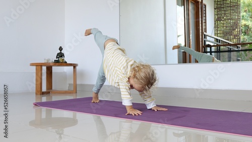 A little blond girl of about three is learning to do an asana on a yoga mat. She got into a dog pose and lifts one leg up. Then he sits down and looks at his mother's approving look. photo