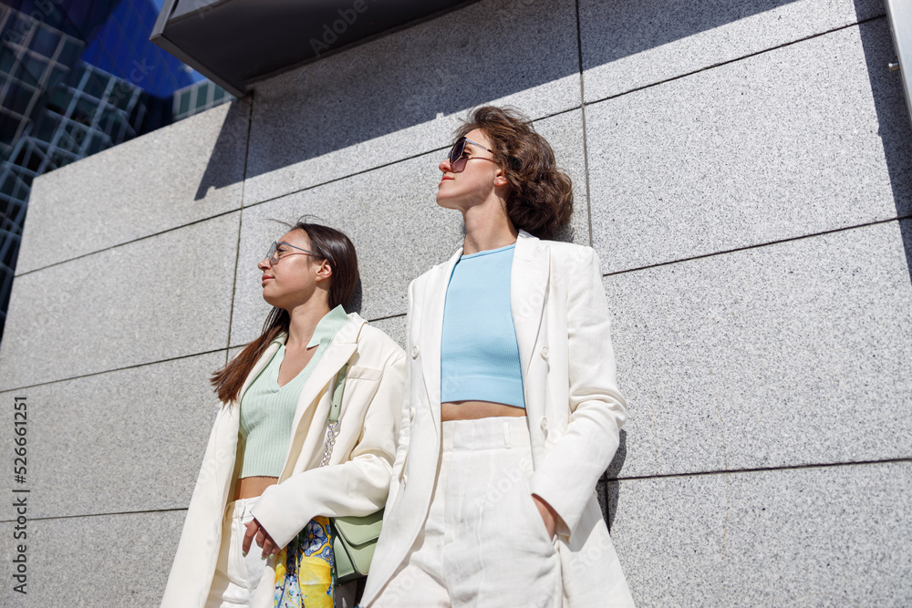 Fototapeta premium Two woman friends have a rest after walking in city standing near wall