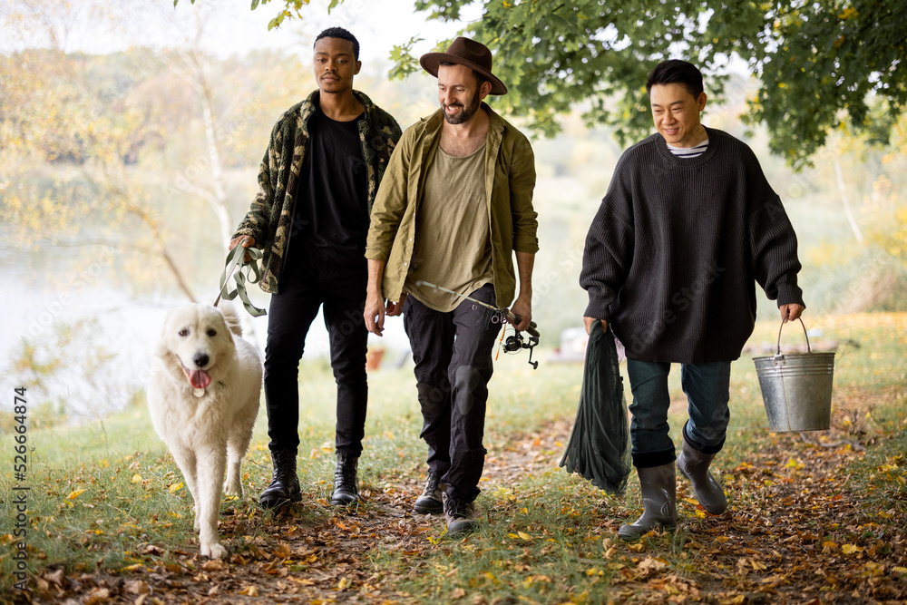 Multiracial male friends walking with Maremmano-Abruzzese Sheepdog and fishing equipment in nature. Concept of weekend and vacation in nature. Idea of friendship. Men between forest and river