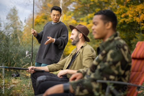 Asian man talking to male multiracial friends during fishing in nature. Men resting and spending time together on river or lake shore. Concept of leisure, hobby and weekend in nature. Friendship