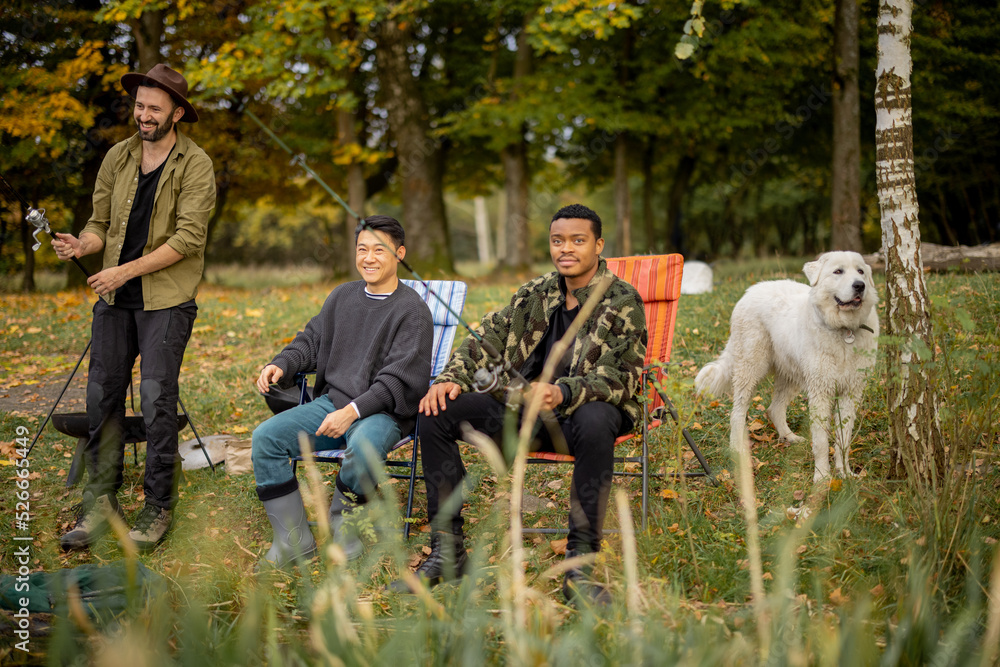 Fototapeta premium Pleased multiracial male friends resting and fishing on river or lake coast. Concept of leisure, hobby and weekend in nature. Idea of friendship and spending time together. View from the waterside