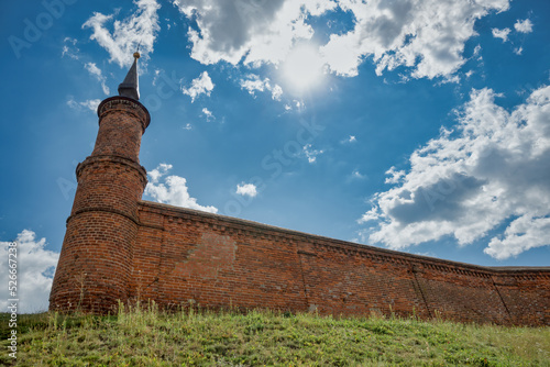 Part of a brick fortress wall with a tower against a blue cloudy sky and the sun.