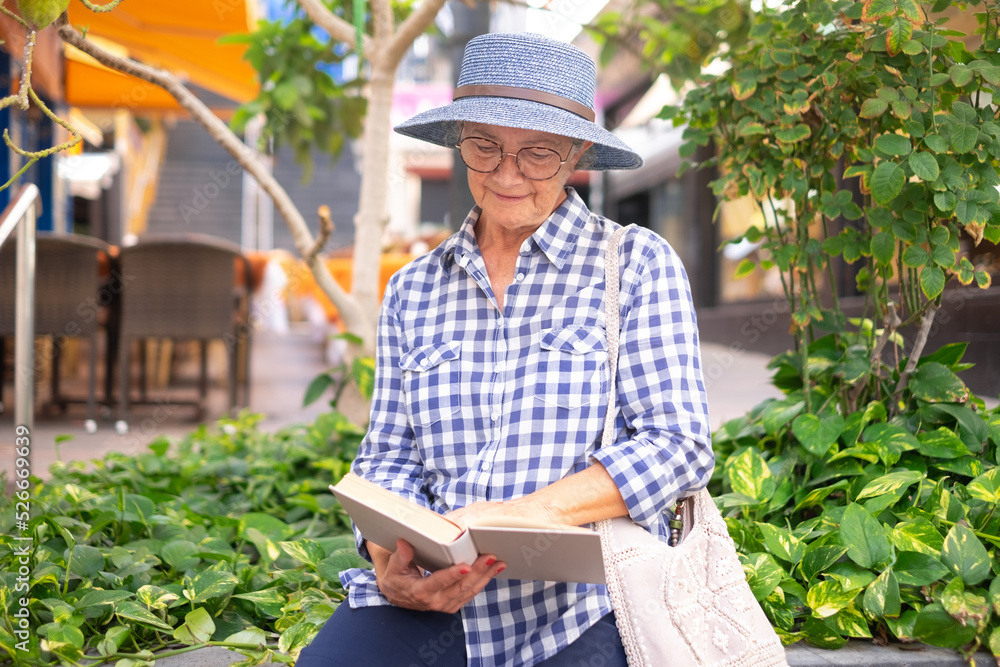 Attractive senior woman sitting in the city holding a book in her hand - caucasian lady enjoying free time and retirement Attractive senior woman sitting in the city holding a book in her hand - caucasian lady enjoying free time and retirement