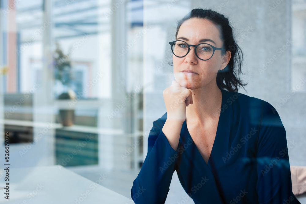Businesswoman sitting with hand on chin seen through glass Businesswoman sitting with hand on chin seen through glass