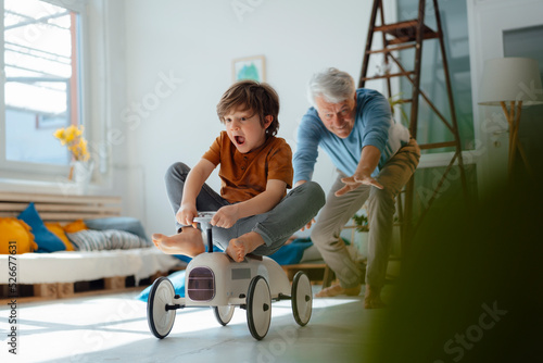 Cheerful grandfather playing with grandson sitting on toy car in living room photo