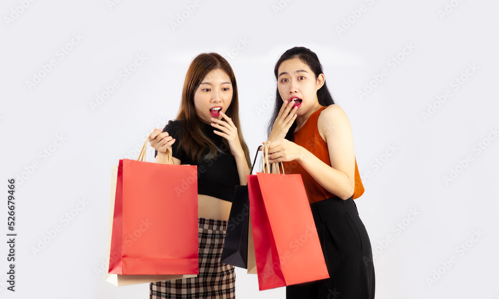 Two asian women in casual costume carrying red black and brown paper shopping bags posing shock and surprised on white background. Two asian women in casual costume carrying red black and brown paper shopping bags posing shock and surprised on white background.