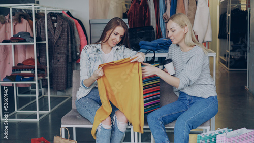 Cheerful female customer showing her friend purchased yellow jumper while sitting together in luxurious boutique. Girls are excited about price and quality of clothing. © silverkblack