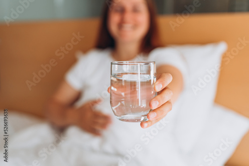 Pregnant woman with glass of water sitting on bed at home. Millennial Caucasian woman wake up in cozy white bed in modern apartment in the morning. Concept of healthy lifestyle, selective focus