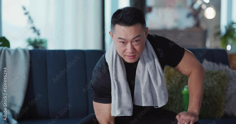 Portrait of Handsome Strong Japanese Young Man in Sportswear Sitting on Sofa in Living Room Lifting Heavy Black Dumbbell SMiling to Camera. Portrait of Handsome Strong Japanese Young Man in Sportswear Sitting on Sofa in Living Room Lifting Heavy Black Dumbbell SMiling to Camera.