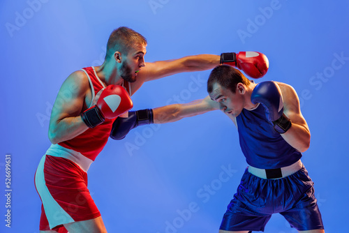 Studio shot of male professional boxers in red and blue sports uniform boxing isolated on blue background in neon light. Sport, skills, power, training, energy © master1305