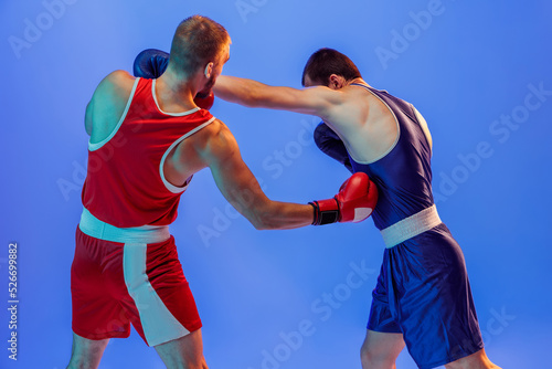 Left hook. Male professional boxers in red and blue sports uniform boxing isolated on blue background in neon light. Sport, skills, power, training, energy