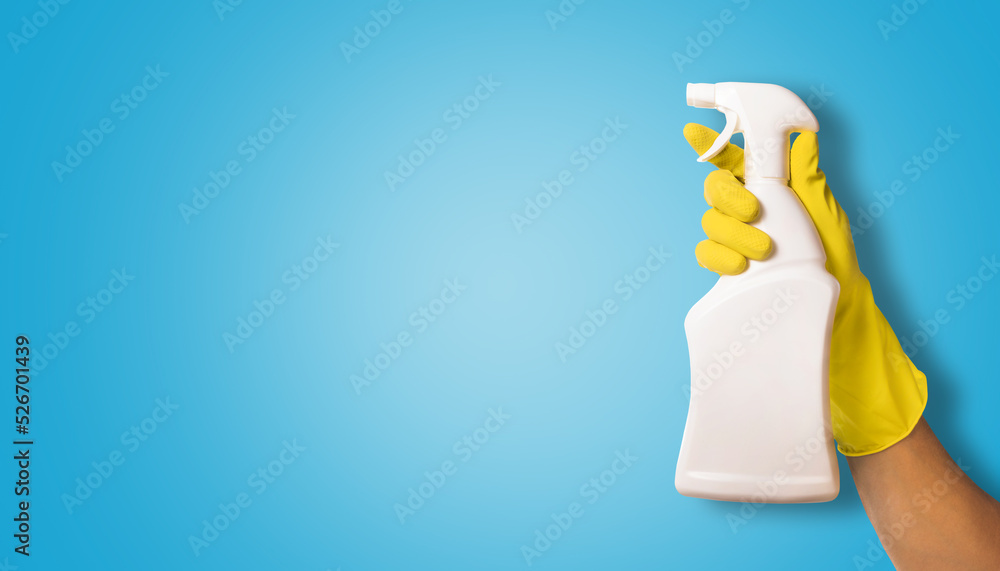 hand in yellow rubber glove holding white blank cleaning spray bottle on blue background. copy space - obrazy, fototapety, plakaty 