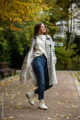 European elegant young woman in a stylish trench coat in a white sweater and blue jeans walking in a park. warm autumn day. © Mykola