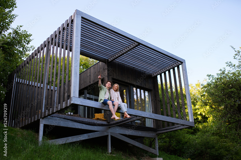 Young couple sitting and cuddling in hammock terrace in their new home in tiny house in woods, sustainable living concept. - obrazy, fototapety, plakaty 