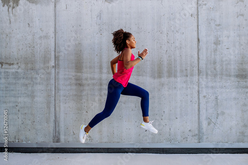 Young multiracial girl jogging and jumping in city, in front of concrete wall, side view.