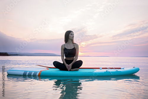 Young surfer girl meditating on surfboard on the lake at sunrise. © Halfpoint