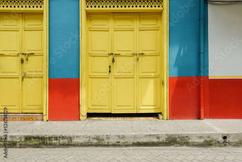 colorful facade of a traditional house in a colombian village. Two large closed yellow gates in a blue and red wall. An empty scene with sidewalk. Looks caribian and mediterranean.  photo