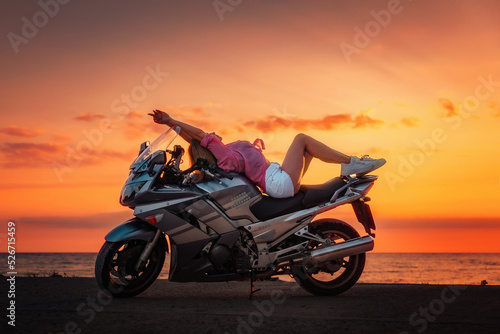 Freedom. Sexy young woman posing on the motorcycle. Golden sunset and motorbike on the background. Side view. Concept of World Motorcyclist Day and moto trips