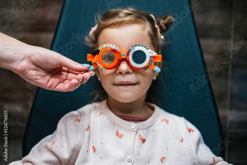Beautiful and adorable little girl with optometry glasses receiving ophthalmology treatment. Doctor ophthalmologist checking her eyesight with modern equipment. Focus on glasses.