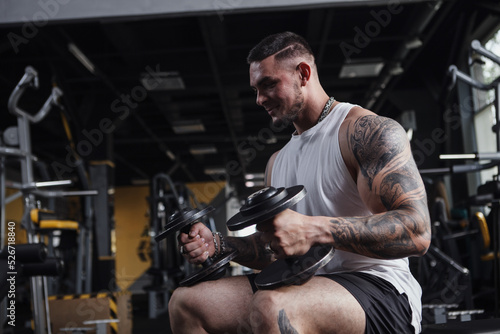 Handsome tattooed muscular bodybuilder holding heavy dumbbells, sitting at the gym