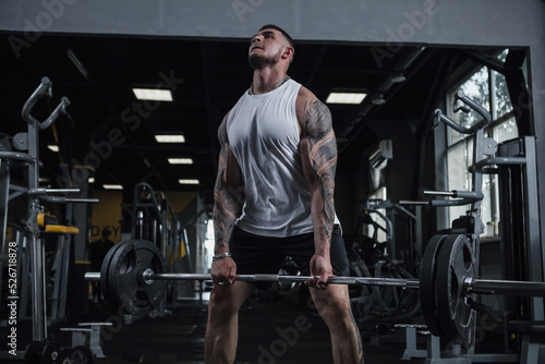 Huge muscular tattooed bodybuilder lifting heavy barbell at gym