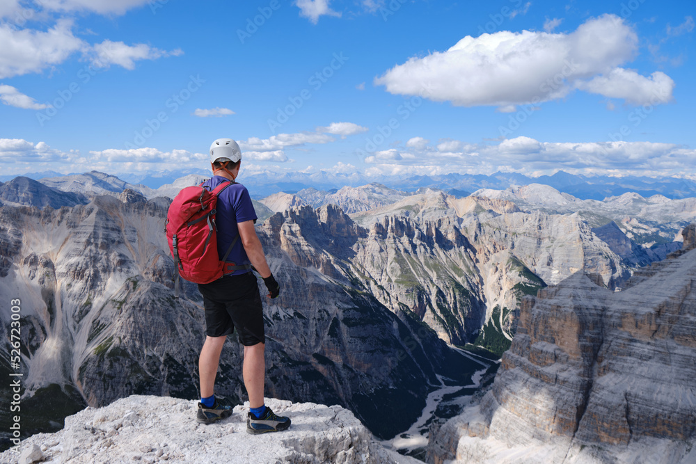 Man hiker with helmet and red backpack at the edge of a cliff in the Dolomites mountains, Italy. Summer, active, activities, mountaineering, copy space.