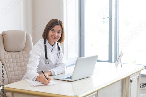 Portrait photo of young and beautiful smiling female doctor in white medical gown sitting and working on laptop in the office of the modern clinic. Online consultation and distant cure concept
