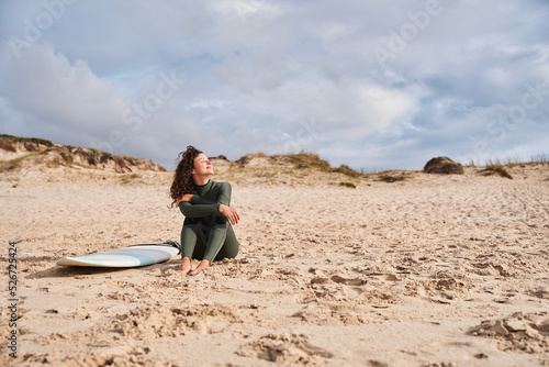 Calm girl surfer looking at the nature while sitting at the sandy beach with surf board