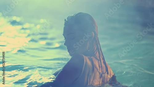 Beautiful woman posing in pool water under neon color light. Party, attractive chick in elegant feminine gypsy style dress enjoying night time. Hippie lifestyle, bohemian clothing trend, fashion.