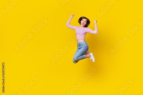 Full length photo of girlish adorable pretty woman dressed striped top jeans sneakers jumping hands up isolated on yellow color background