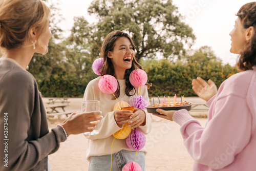 Three happy young caucasian women celebrate birthday outdoors in summer. Brunettes laugh and enjoy day, wear sweaters. Party concept