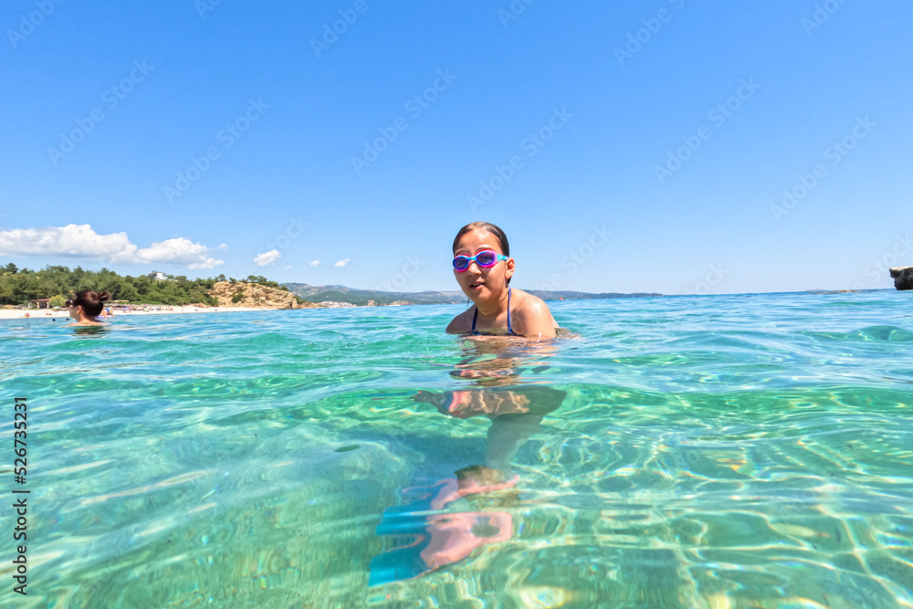 Summer vacation portrait of child girl with swimming goggles in transparent turquoise color sea water. Clear blue sky. 