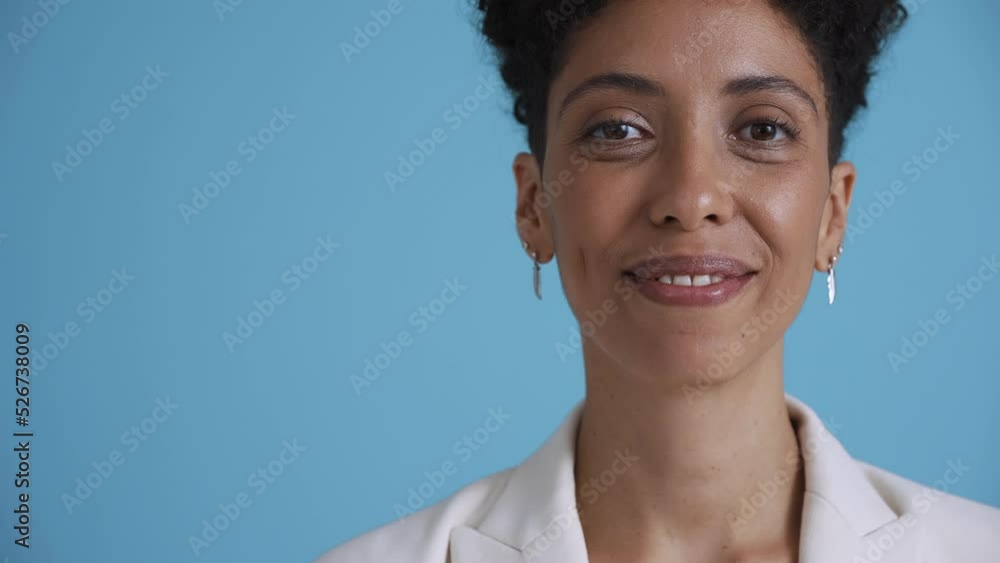 Face of smiling hispanic curly haired woman wearing white jacket looking at the camera in the blue studio Face of smiling hispanic curly haired woman wearing white jacket looking at the camera in the blue studio