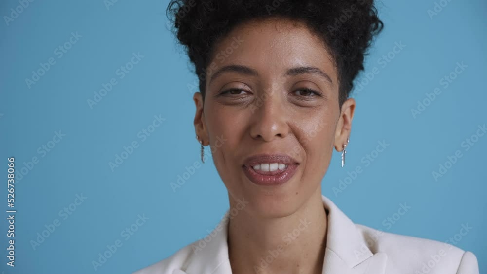 Face of happy hispanic curly haired woman looking around and showing shh gesture at the camera in the blue studio Face of happy hispanic curly haired woman looking around and showing shh gesture at the camera in the blue studio