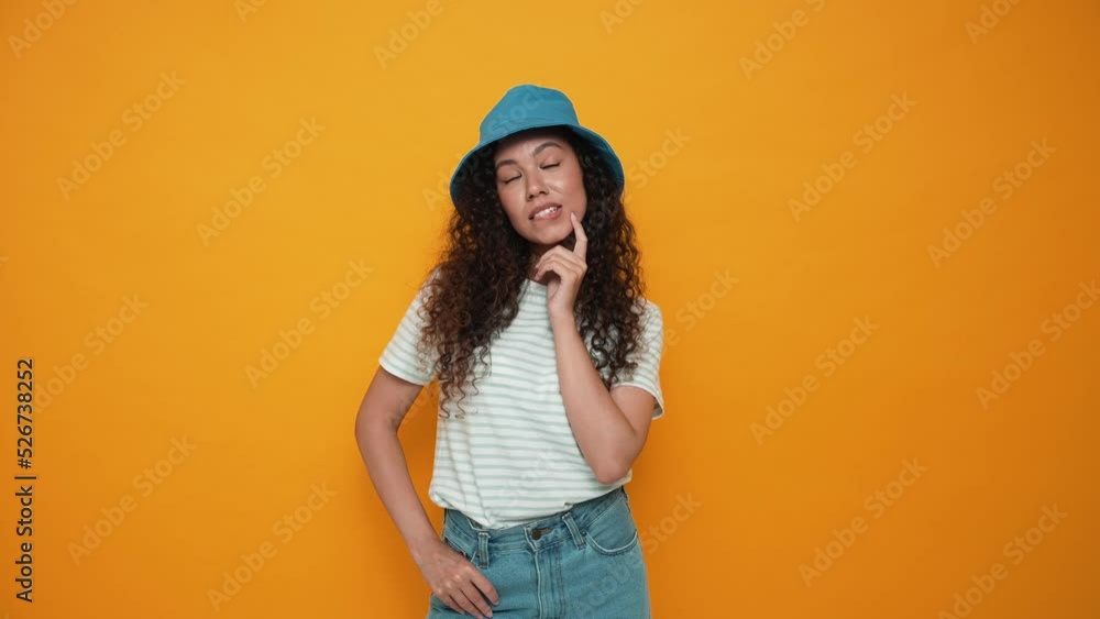 Positive brunette curly haired woman wearing panama hat thinking about something in the yellow studio Positive brunette curly haired woman wearing panama hat thinking about something in the yellow studio