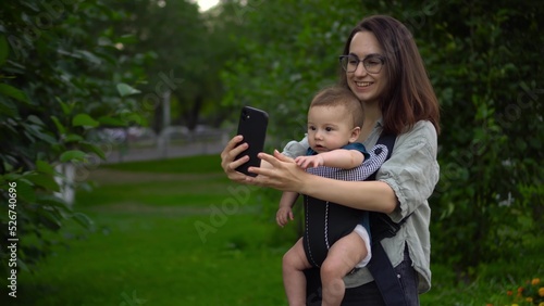 A young mother walks in the park with her little son and makes a video call on the phone. A girl in glasses walks with a newborn. A child in a kangaroo-backpack.