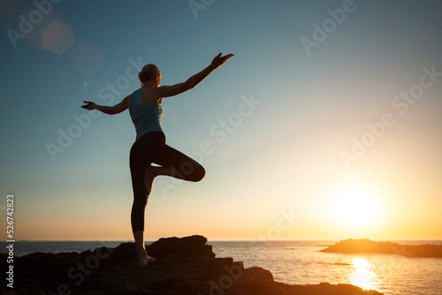 A woman does yoga, doing asana on the oceanfront during sunset.