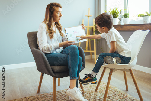 Professional female child psychologist working with a little preschool boy in a bright office. Woman makes notes about the condition of the boy on paper. Children's mental therapy. photo