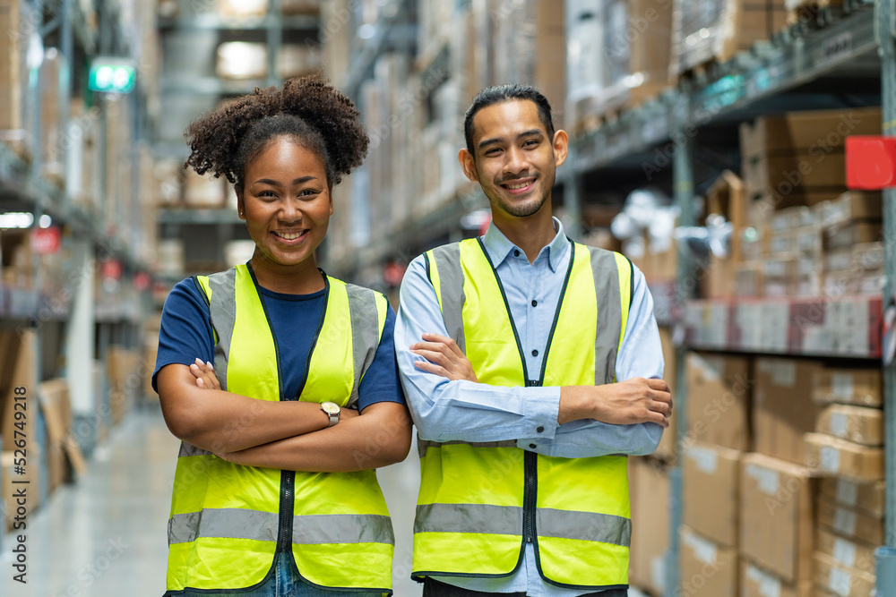Naklejka premium Asian male and African American female employees wearing colleague uniforms standing with their arms crossed and proud of warehouse and freight service.