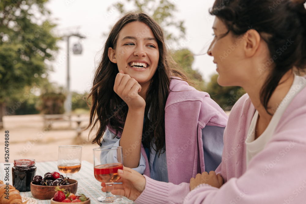 Two cheerful young caucasian women with booze are having picnic outside city spring. Brunettes wear sweatshirts in warm weather. Vacation concept.