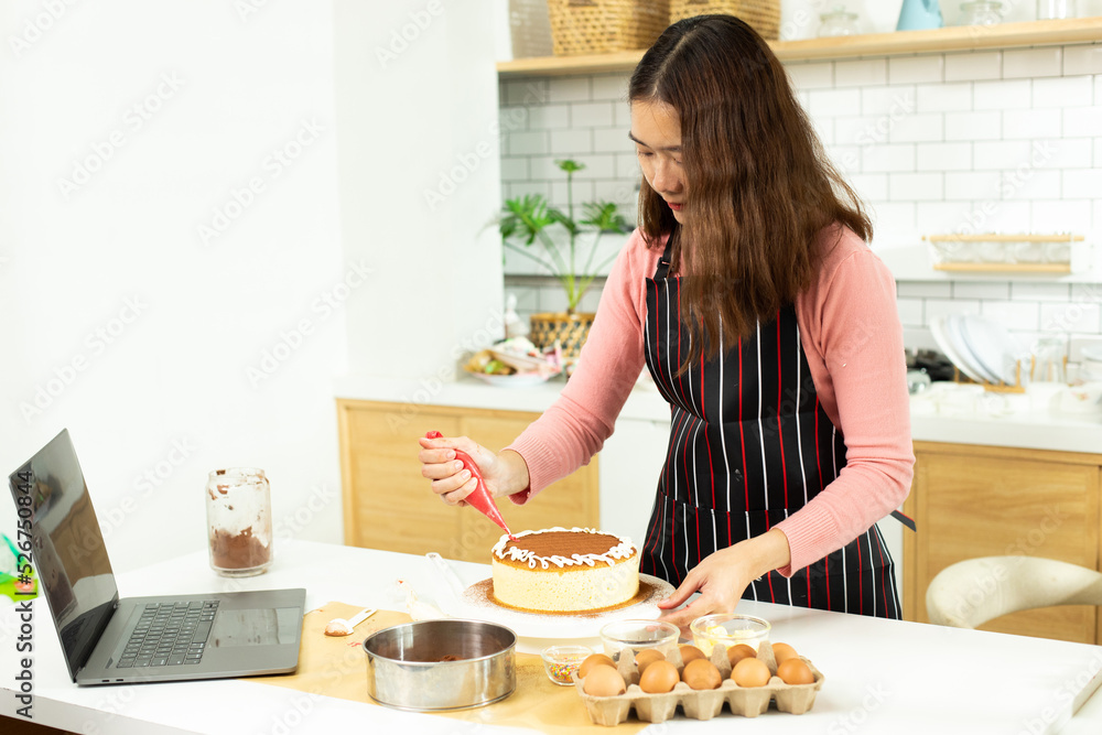 Asian young adult woman learn how to bakery cooking cake online course in kitchen. Female mother video to cyberspace class on laptop as blogger social media content and streaming learning, copy space