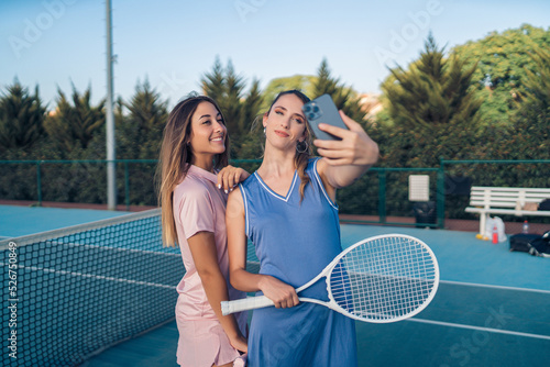 Dos chicas vestidas con ropa de tenis rosa y morada paseando juntas y haciendose selfies con smartphone en cancha de tenis
