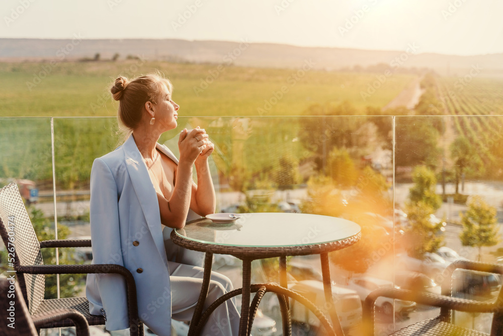 A middle-aged woman sits in a street cafe overlooking the mountains at sunset. She is dressed in a blue jacket and drinks coffee admiring the nature. Travel and vacation concept. A middle-aged woman sits in a street cafe overlooking the mountains at sunset. She is dressed in a blue jacket and drinks coffee admiring the nature. Travel and vacation concept.