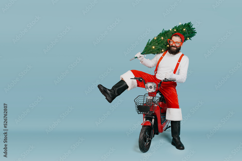 Santa in a red hat, big heart-shaped glasses, pants with suspenders sits on an electric scooter putting a Christmas tree with a garland on his shoulder on a blue isolated background with copy space Santa in a red hat, big heart-shaped glasses, pants with suspenders sits on an electric scooter putting a Christmas tree with a garland on his shoulder on a blue isolated background with copy space