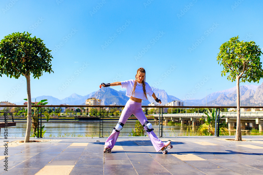Obraz premium Portrait of beautiful blonde teenager girl with cool pigtails , white t-shirt and flared pink pants skating on rollers in park